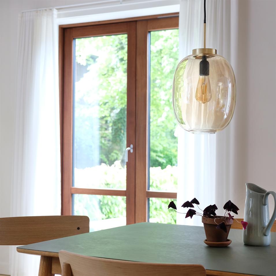 En klar glaspendel hænger over et grønt bord; et lille potteplante og en keramikkande står på bordet foran en trædør med hvide gardiner og grønt haveudsyn.