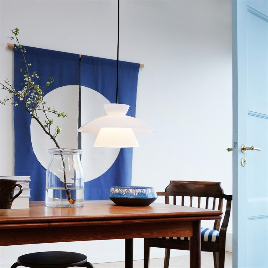 En hvid pendel hænger over et træspisebord; på bordet står en glasvase med blomstrende kvist og en blå mønstret skål, foran et blåt vægtæppe med hvid cirkel og en åben lyseblå dør.