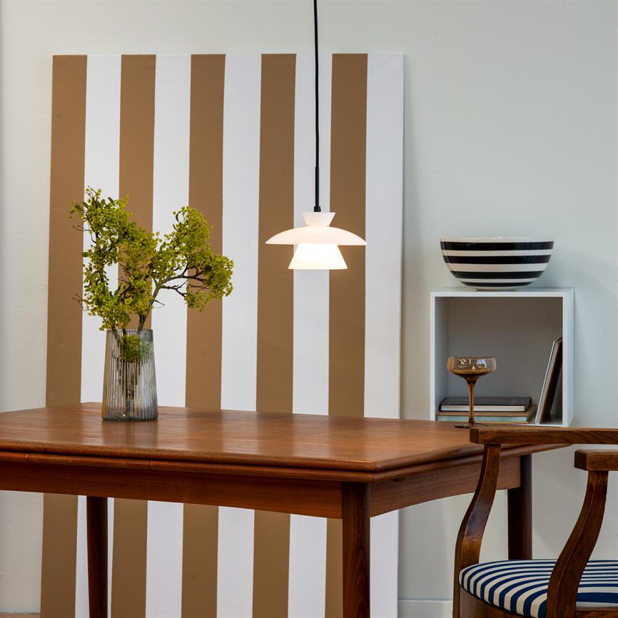 Et træbord bærer en klar glasvase med grønne grene, mens en hvid pendellampe hænger og lyser; bag bordet står et lodret brun-hvidt stribet panel, hylde med skål og stol.