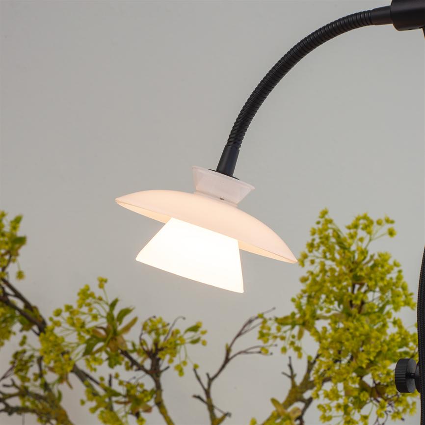 En hvid to-lags lampeskærm på en sort fleksibel arm lyser nedad; den kaster varmt lys foran en lys væg med grønne, blomstrende grene i baggrunden.