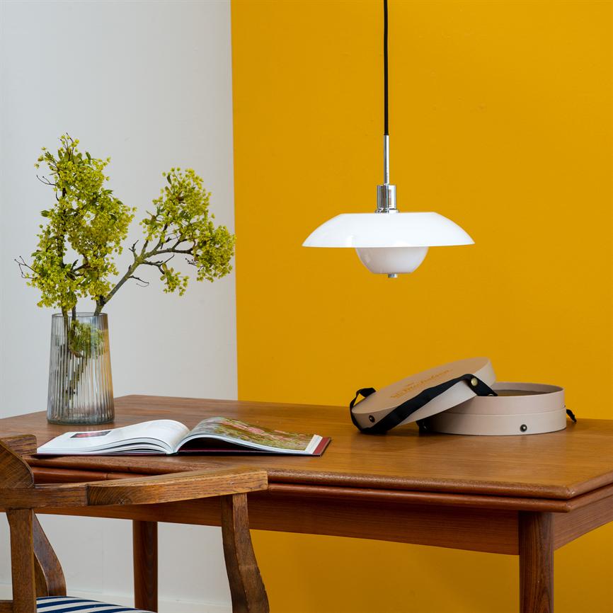 En hvid pendellampe hænger over et træbord; et åbent bogblad, en vase med grønne blomster og en åben rund æske ligger på bordet foran en gul væg. Der er utydelig tekst på æskens låg, ikke læsbar.