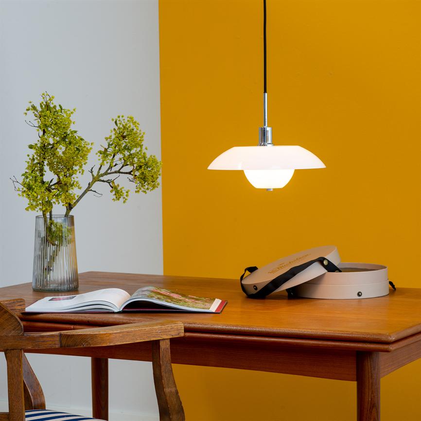 En ophængt hvid pendellampe lyser over et træbord; et åbent magasin, to runde æsker og en glasskål med grønne grene ligger på bordet foran en gul væg.