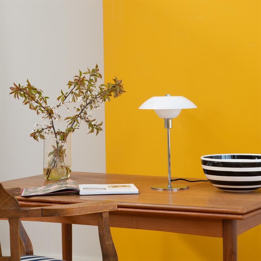 En hvid bordlampe står på et træbord; en glasvase med blomster til venstre, en åben bog foran og en sort-hvid stribet skål til højre; bagvæggen er delt i hvid og gul.