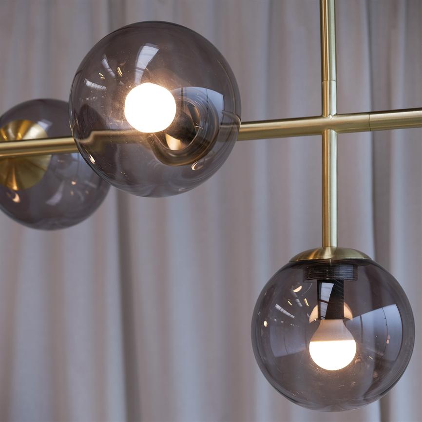 Tre røgfarvede glaskupler med tændte pærer hænger fra en gylden metalramme foran bløde, lyse gardiner i et indendørs rum.