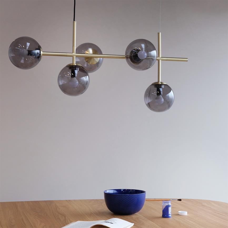 Pendellampe med fem røgfarvede glaskupler hænger over et lyst træbord, hvor en blå skål, en pensel, en åben bog og en lille maledåse står mod en neutral væg.