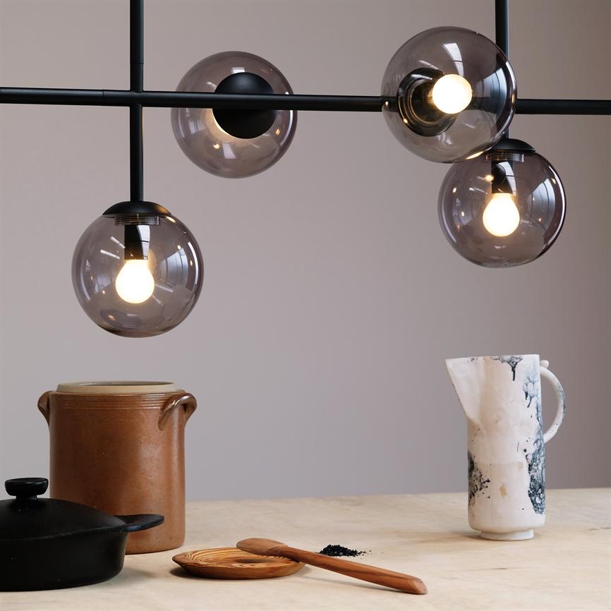 En moderne pendel med fire røgfarvede glaskupler lyser ned over et køkkenbord; under står en brun keramikkrukke, en sort støbejernsgryde, en marmoreret hvid kande, en træske og lidt krydderi.