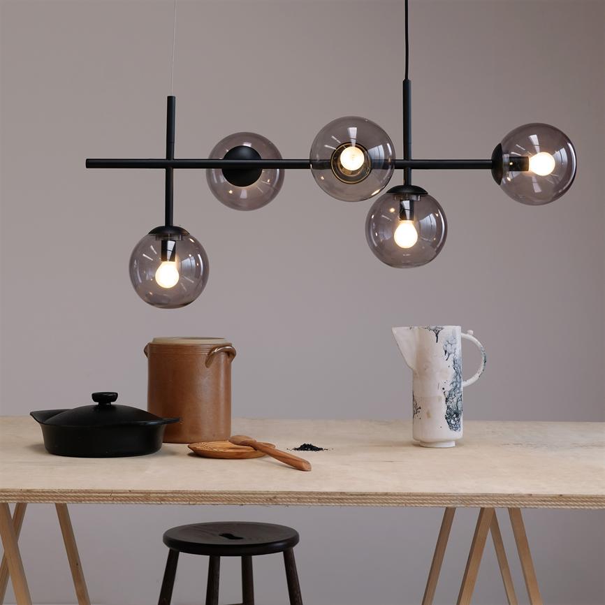 En moderne loftlampe med fem røgfarvede glaskupler hænger og lyser over et lyst træbord med krukke, hvid marmoreret kande, støbejernsgryde og træske i et minimalistisk rum.