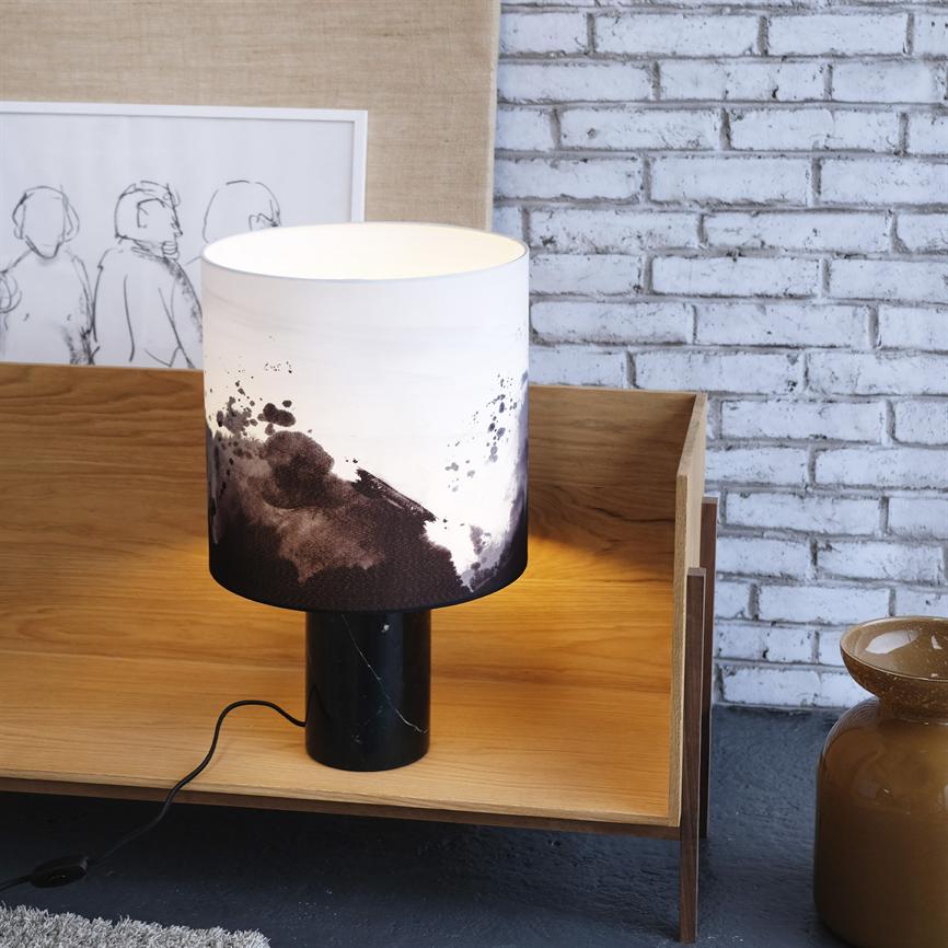 Bordlampe står på en træhylde og udsender varmt lys; hvid skærm med sorte akvarelpletter, sort marmorfod; omgivet af en indrammet skitse på væggen, hvid murstensbaggrund og en brun vase.