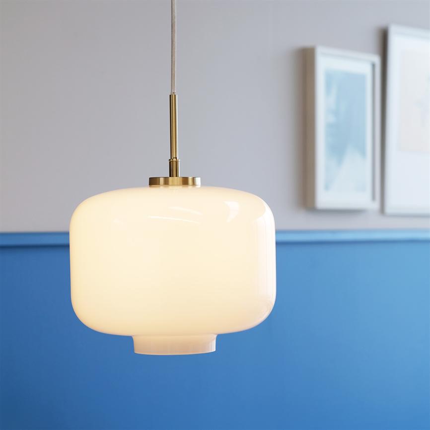 En opalhvid glas-hængelampe lyser blødt, hænger i messingophæng foran en grå væg med blå brystning og to slørede indrammede billeder.