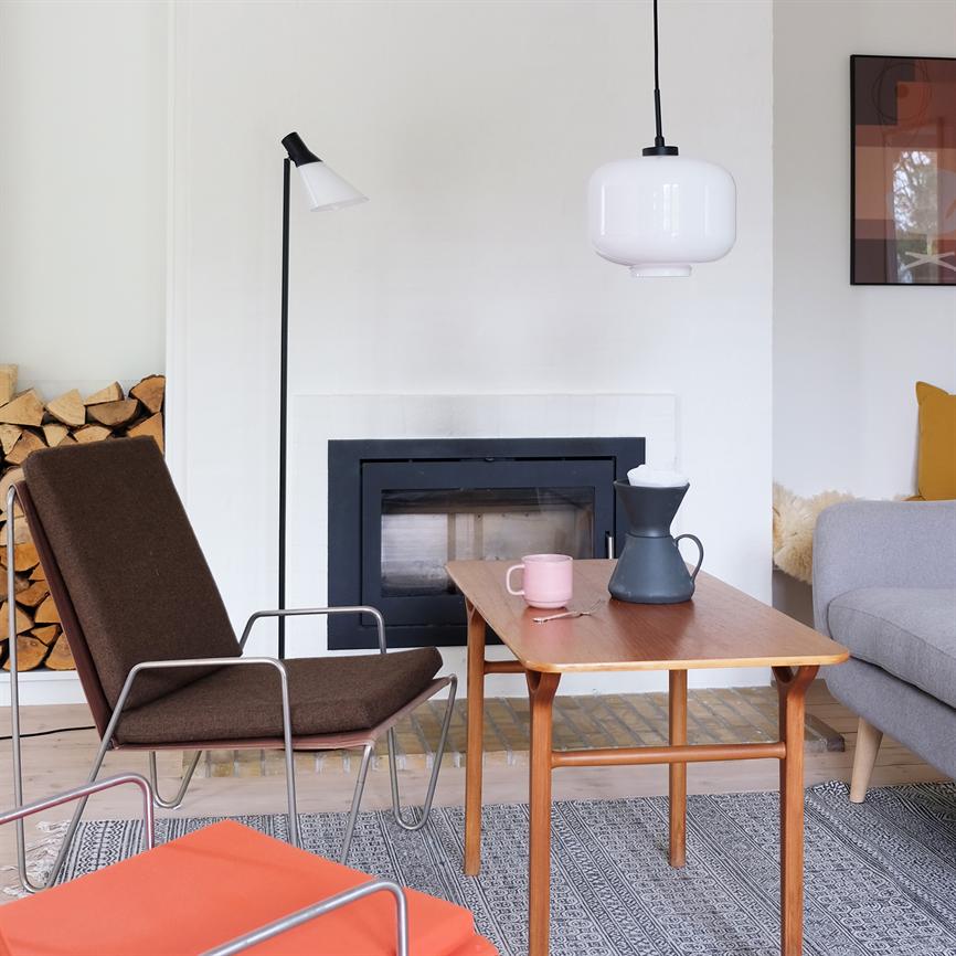 Et teak sofabord står foran en lukket pejs; et lyserødt krus og en sort kaffekande står på bordet. Omgivet af lænestol, grå sofa, brænde og hængelampe.