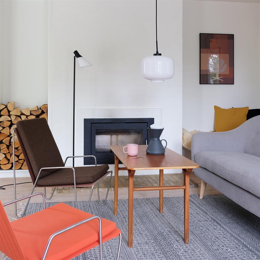 Et træ-sofabord står på et mønstret tæppe foran en lukket pejs; en mørk kande og en lyserød kop står på bordet; moderne stue med sofa, stole og brænde.