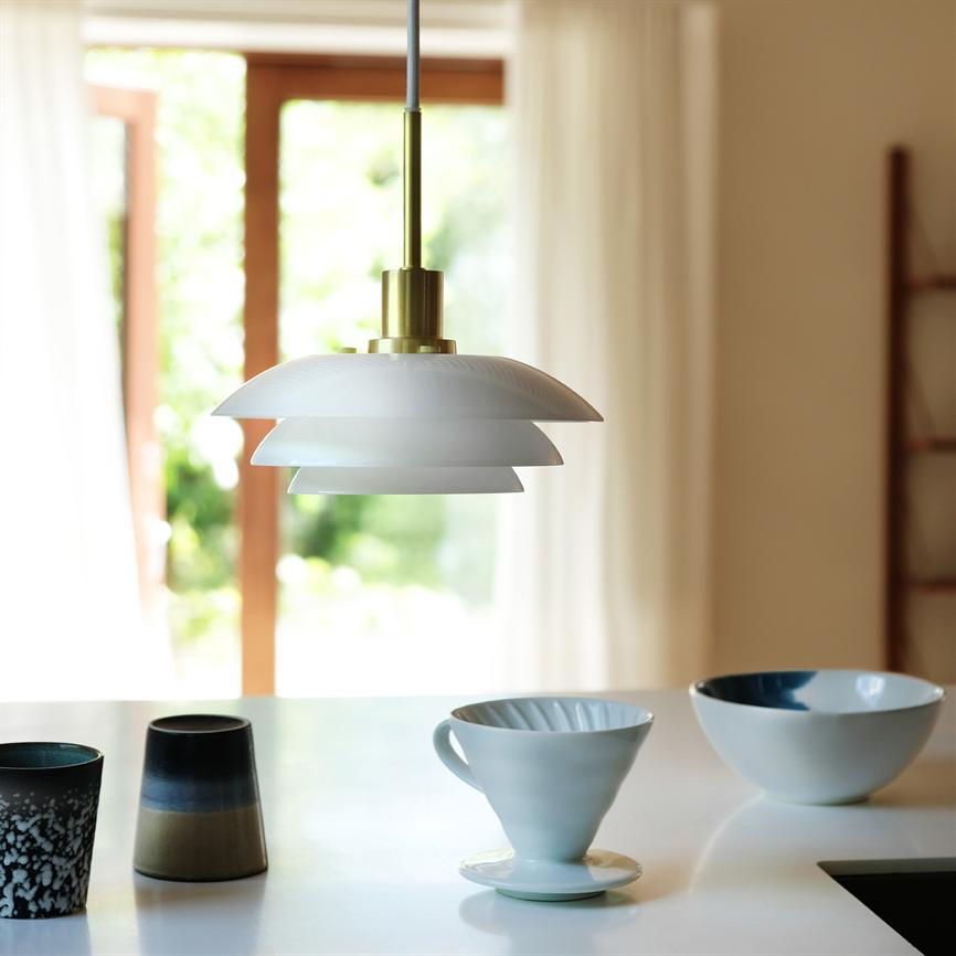 En hvid, lagdelt pendellampe hænger og kaster blødt lys over et køkkenbord med keramikkopper, en kaffetragt og en skål, foran store vinduer med lyse gardiner og grønt udenfor.