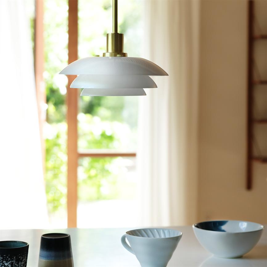 En hvid, lagdelt pendellampe hænger over et bord; under den står keramikkrus og skåle, ved et lyst vindue med bløde gardiner og grønt udsyn udenfor.