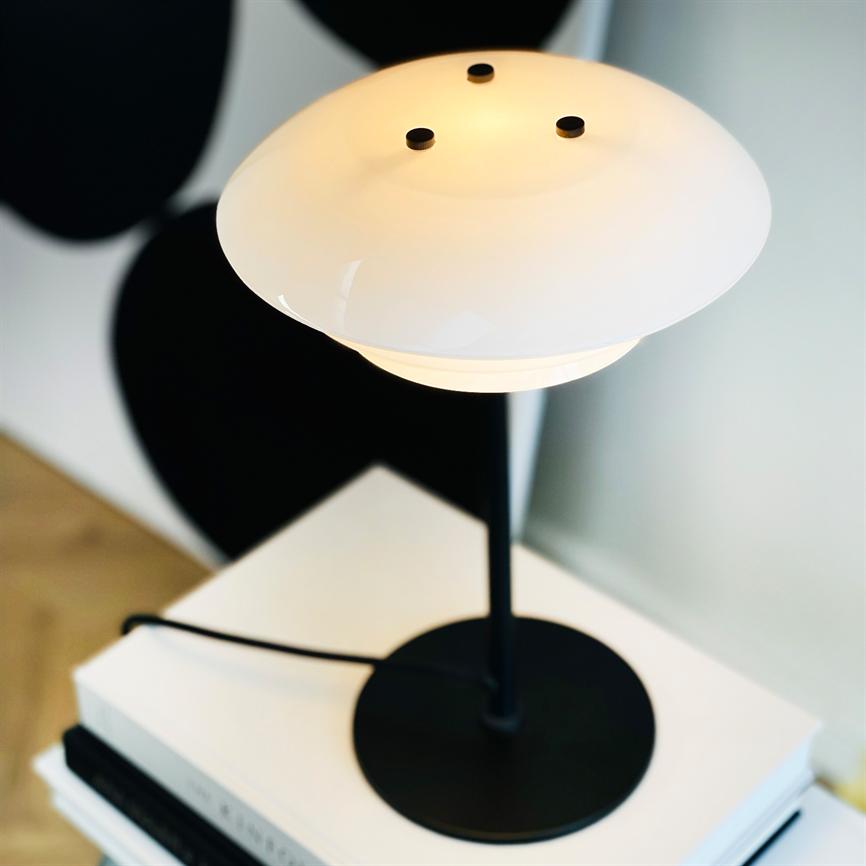 En sort bordlampe med hvid, skålformet skærm lyser blødt, placeret på en stak hvide bøger på et trægulv, med en utydelig sort baggrund.