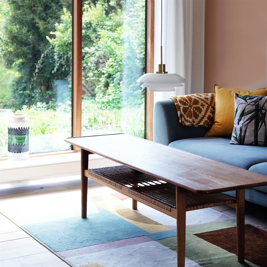 Sofabord i mørkt træ står på et farverigt tæppe foran en blå sofa; over bordet hænger en hvid pendel, og store vinduer åbner mod en frodig have.