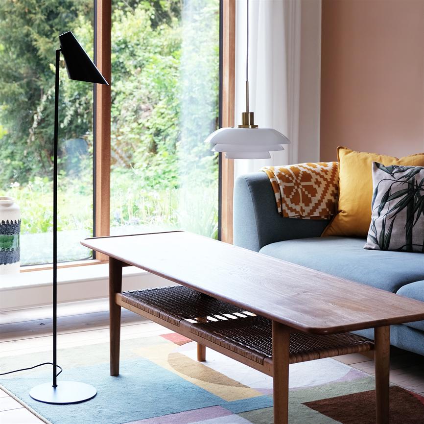 Et sofabord i træ står foran en blå sofa, omgivet af en gul pude, en pendellampe med messingdetalje og en sort gulvlampe ved et stort vindue mod en have.