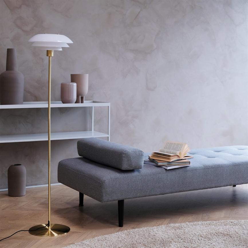 En grå daybed har en åben bog og en stak bøger på sædet; en gulvlampe og hvide hylder med vaser står i et lyst, minimalistisk rum med rå væg.