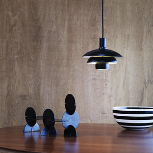 Sort metalpendel hænger over et rundt træbord; tre sammenføjede sorte skiveelementer står på bordet, og en sort-hvid stribet skål står til højre foran en lyst træbeklædt væg.