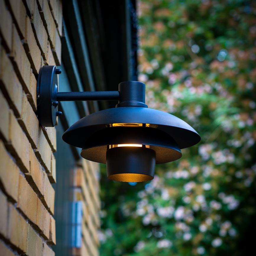 En sort, rund væglampe udsender varmt, nedadrettet lys; den er monteret på en gul murstensvæg udenfor med sløret grønt løv og blomster i baggrunden.