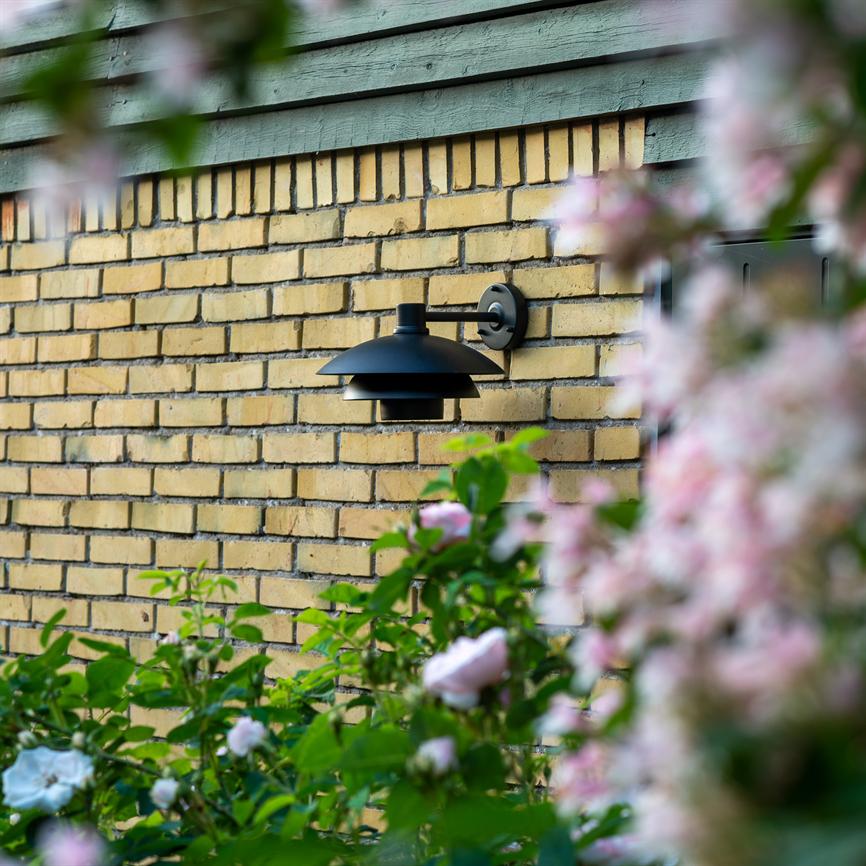 Sort væglampe hænger på en gul murstensfacade, omgivet af grønne buske og bløde lyserøde blomster i forgrunden, under en træbeklædt gesims.