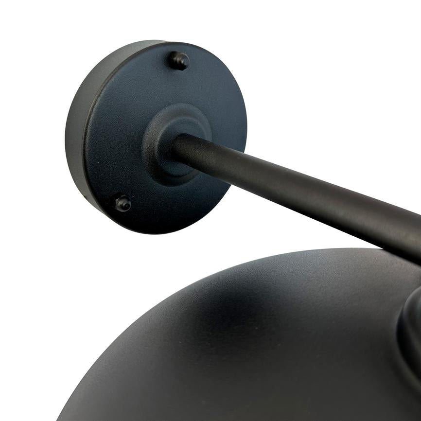 Mat sort metal væglampe: rund vægbeslag og arm strækker sig diagonalt mod en kuppelformet skærm; tæt detalje på en hvid, neutral baggrund.