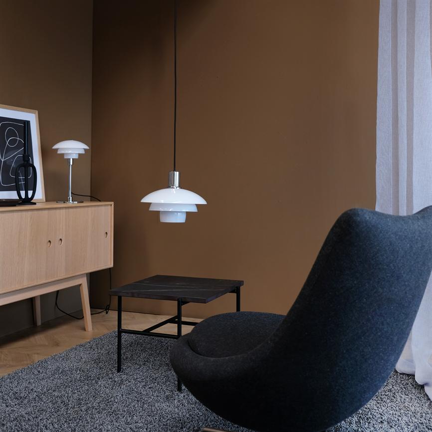 Hvid pendel hænger over et lavt sort sofabord i et minimalistisk stuehjørne med brun væg, lyst egetræs-skab, bordlampe og en mørkegrå kurvet lænestol.