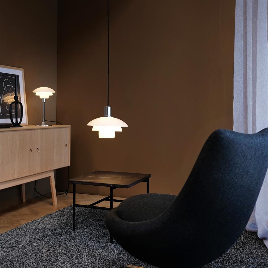 Hængende lampe lyser blødt over et lille sofabord; omgivet af en sort, kurvet lænestol, gråt tæppe, lyst træskab og mørk væg ved et lyst gardin.