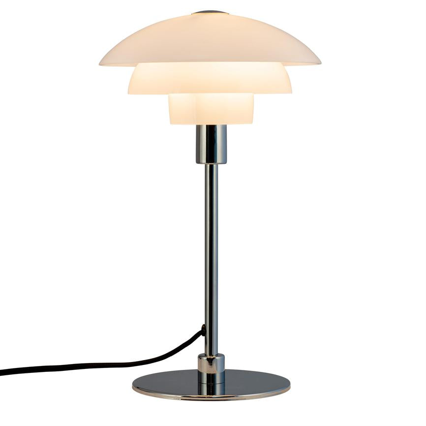 Bordlampe med hvid, tredelt skærm lyser blødt; monteret på slank kromstang og rund kromfod, sort ledning strækker ud til venstre på neutral hvid baggrund.