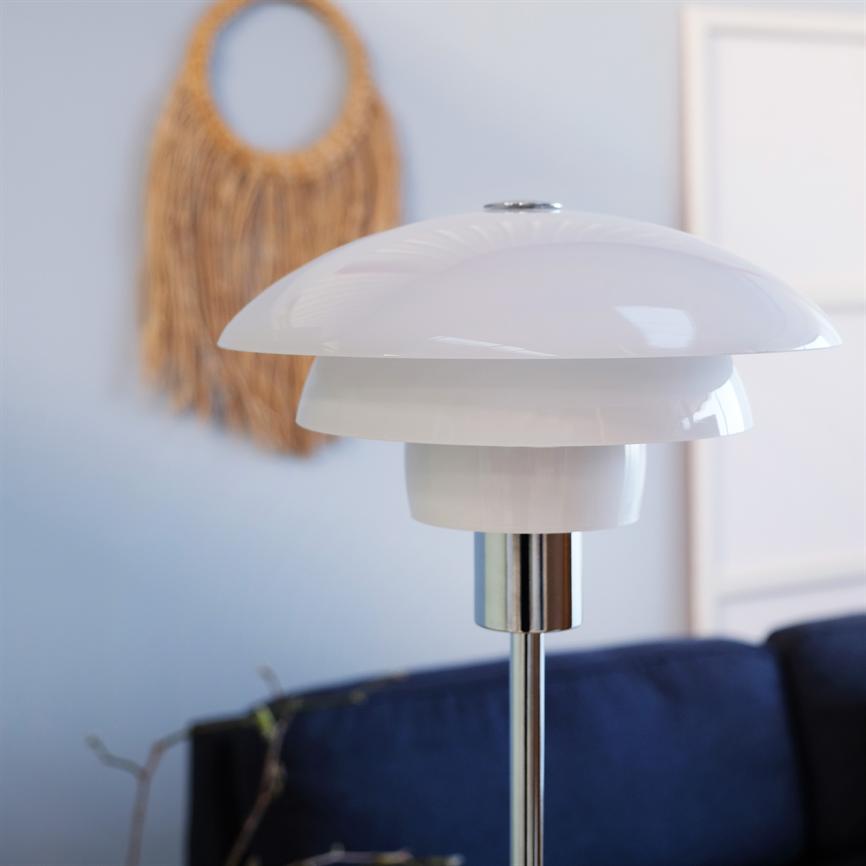 Hvid, blank bordlampe står slukket på en kromstang foran en mørkeblå sofa i et lyst rum med en flettet rattanvægdekoration og en slank billedramme på væggen.