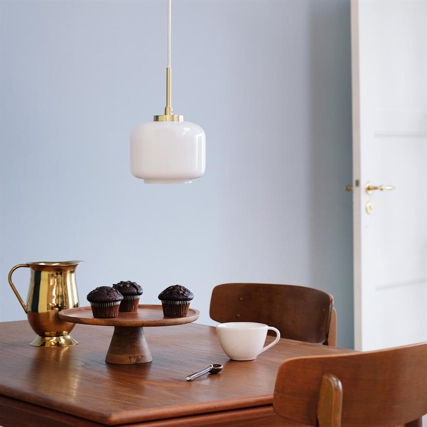 Hvid glaspendel hænger over et træbord; på bordet står et kagefad med tre chokolademuffins, en guldkande og en kop i en enkel spisestue med lyseblå væg og åben dør.