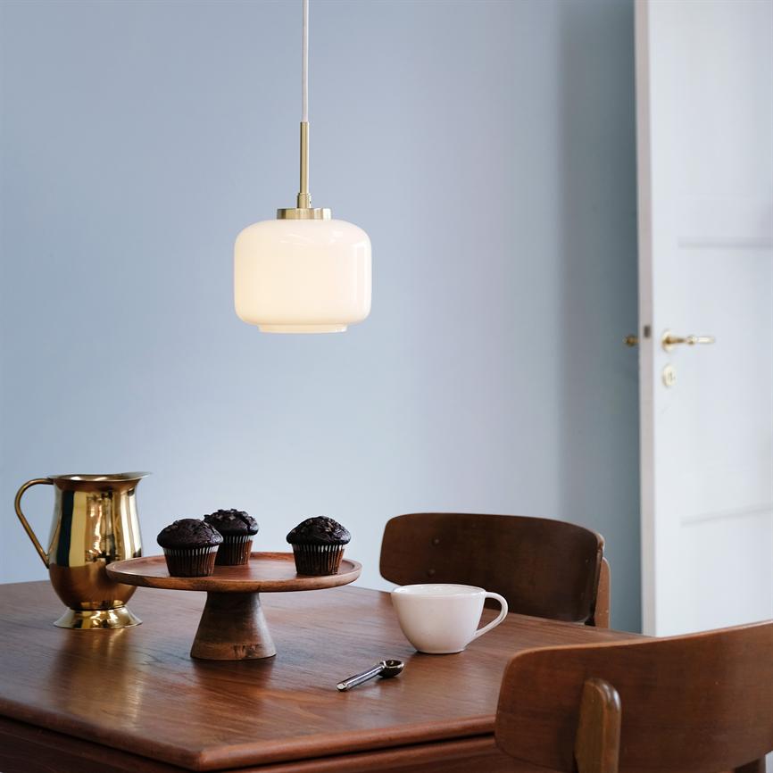 En hvid glaspendel hænger og lyser over et træbord med tre chokolademuffins på en trækageopsats, en hvid kop, en gylden kande og træstole foran en lyseblå væg og åben dør.