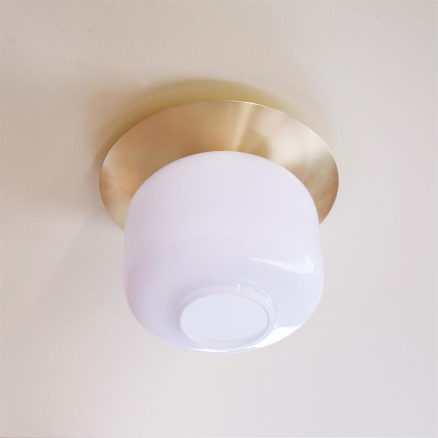 Rund loftlampe i hvidt opalglas, monteret i et lyst loft; den har en mat guldfarvet metalbase og bløde, diffuse reflekser.