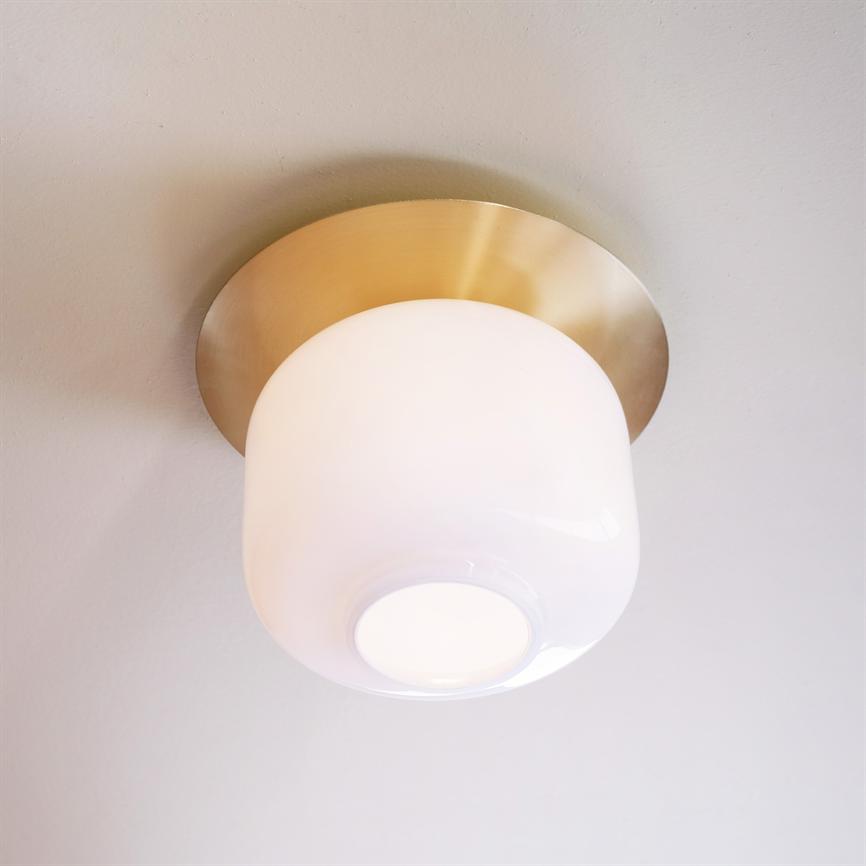 Loftlampe hænger tændt, hvid opalglas-skærm monteret på rund, børstet messingbase; blødt, varmt lys spredes mod et neutralt, lyst loft.