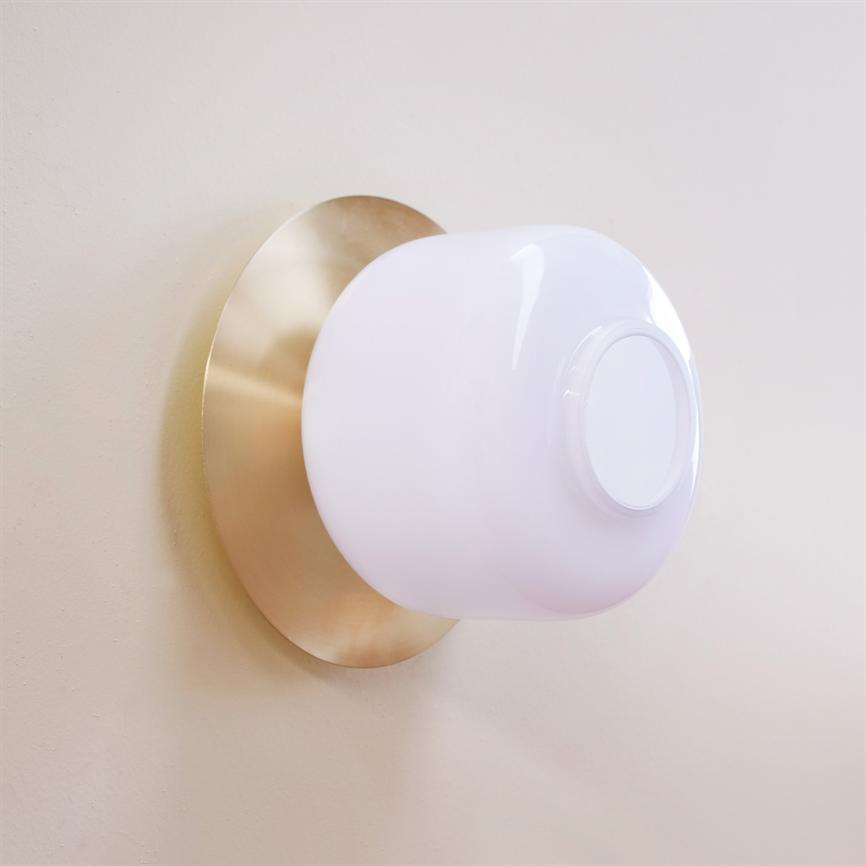 Hvid afrundet væglampe sidder monteret på en rund, børstet messingplade mod en lys beige væg, med bløde reflekser på glasoverfladen i et enkelt, moderne interiør.