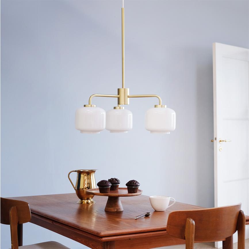 En trearmet messingpendel hænger over et træspisebord; på bordet står en kagedisk med tre chokolademuffins, en gylden kande, en hvid kaffekop og to træstole, lyse vægge og åben dør.