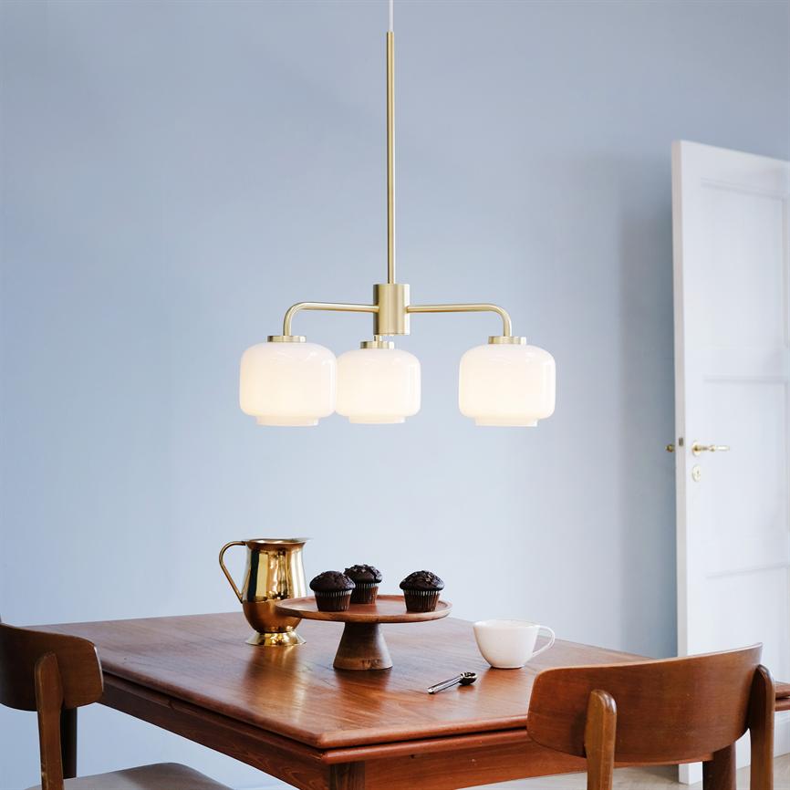 En trearmet loftslampe hænger og lyser over et træspisebord; bordet bærer et kagefad med muffins, en messingkande, kop og ske i en lys, minimalistisk stue.