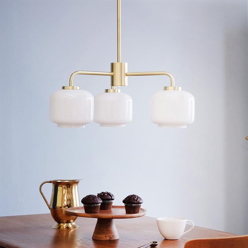 En moderne messingloftlampe med tre hvide glaskuplere hænger over et træbord; på bordet står en guldbelagt kande, et kagefad med tre chokolademuffins og en hvid kop.