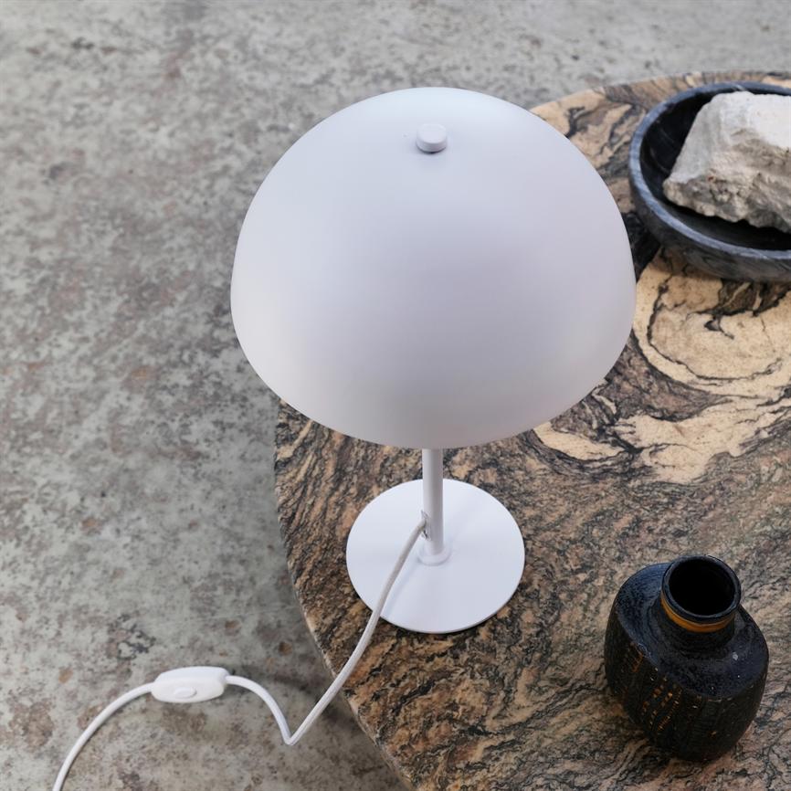 En hvid bordlampe står slukket på et rundt marmorbord ved siden af en mørk keramisk vase og en skål med sten, over et råt betongulv.