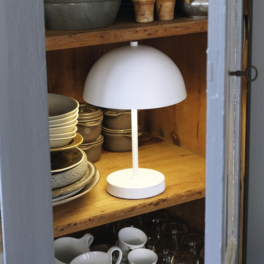 Hvid bordlampe lyser og står på en træhylde i et åbent køkkenskab, omgivet af stakkede tallerkener, skåle og kopper; grå skabslåger ses i forkant.