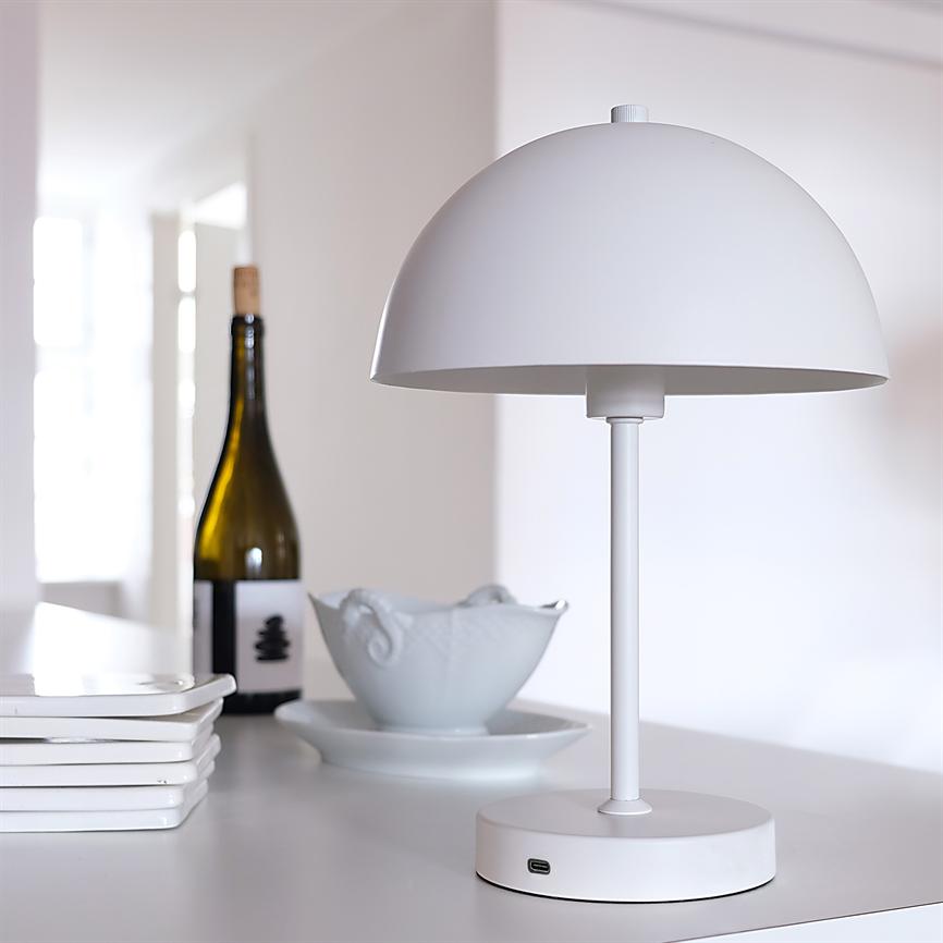 En hvid, halvkugleformet bordlampe står på en hvid bordplade med synlig USB-port; den flankeres af en vinflaske, en keramikskål og en stak hvide tallerkener i et lyst, minimalistisk rum.