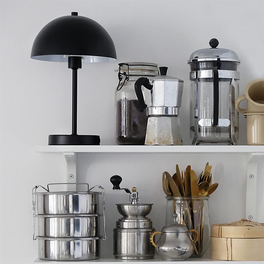 Sort bordlampe står til venstre; en mokkakande, en fransk presse og et glas med kaffe står på øverste hvide hylde; under dem rustfri beholdere, kaffekværn og bestik.