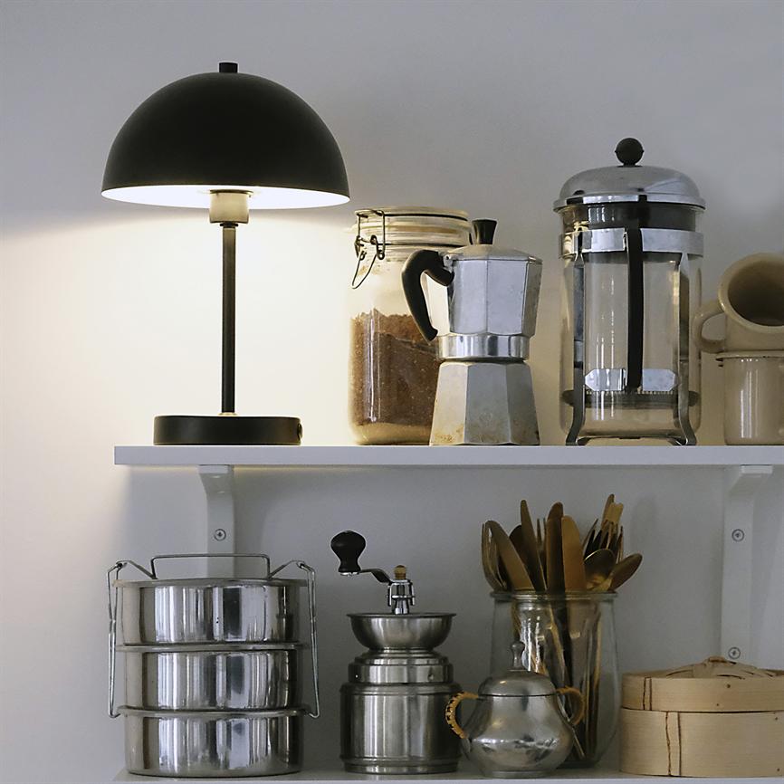 Sort bordlampe lyser en hvid køkkenhylde med kaffetilbehør: glas med formalet kaffe, mokkakande, fransk presse, manuel kværn, metalbeholdere og et glas med træredskaber.