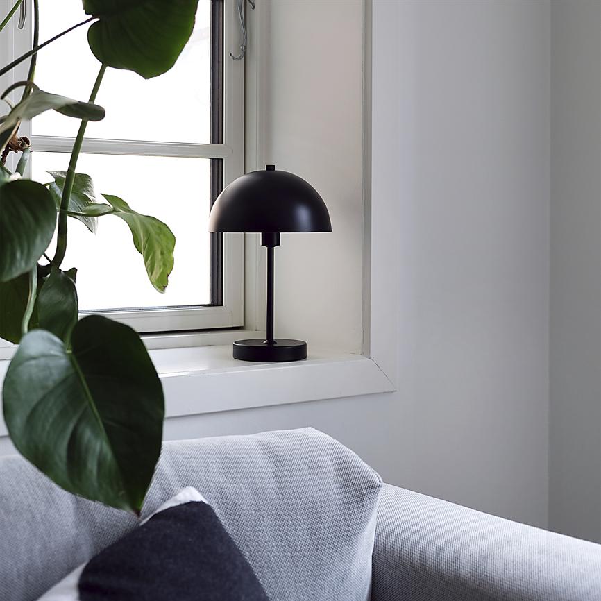 Sort bordlampe står slukket på vindueskarmen; grønne planteblade hænger ind mod vinduet, og en lysegrå sofa med en mørk pude ses i et lyst, minimalistisk rum.
