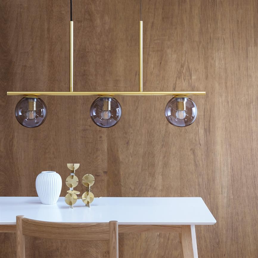 En guldbelagt pendellampe med tre røgfarvede glaskupler hænger over et hvidt bord; foran en træbeklædt væg, med en hvid vase, guldfarvet skulptur og træstol.