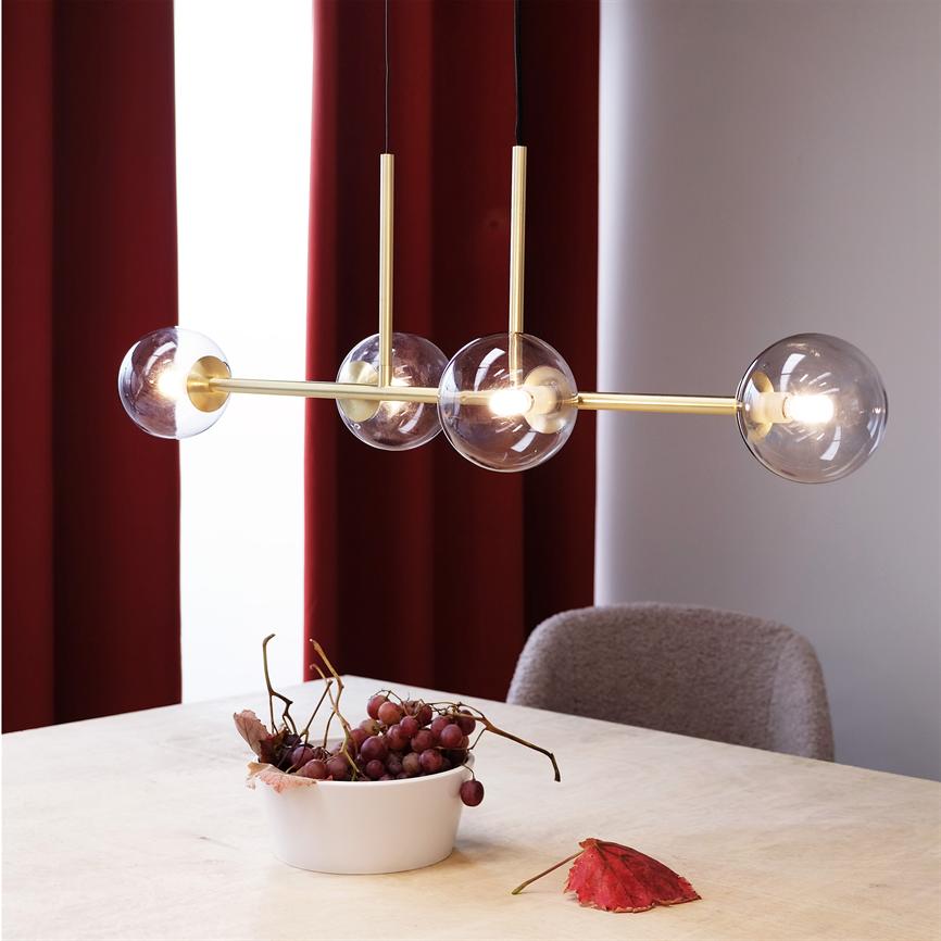 Hængende, moderne lampe med fire glaskupler lyser over et lyst træbord med en hvid skål fyldt med druer; røde gardiner og en polstret stol i baggrunden.