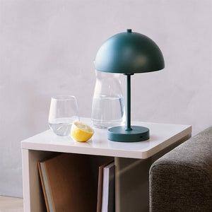 En grøn bordlampe står på et hvidt sidebord; en glas-karaffel, et vandglas og en halv citron ligger ved siden af; bordet står ved en grå sofa foran en lys væg.
