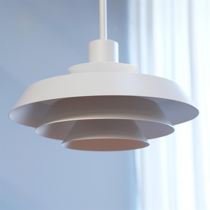 En hvid, lagdelt pendellampe hænger stille fra loftet; dens cirkulære skærme overlapper hinanden. Baggrunden viser en blød blå væg og lyse, gennemskinnelige hvide gardiner.