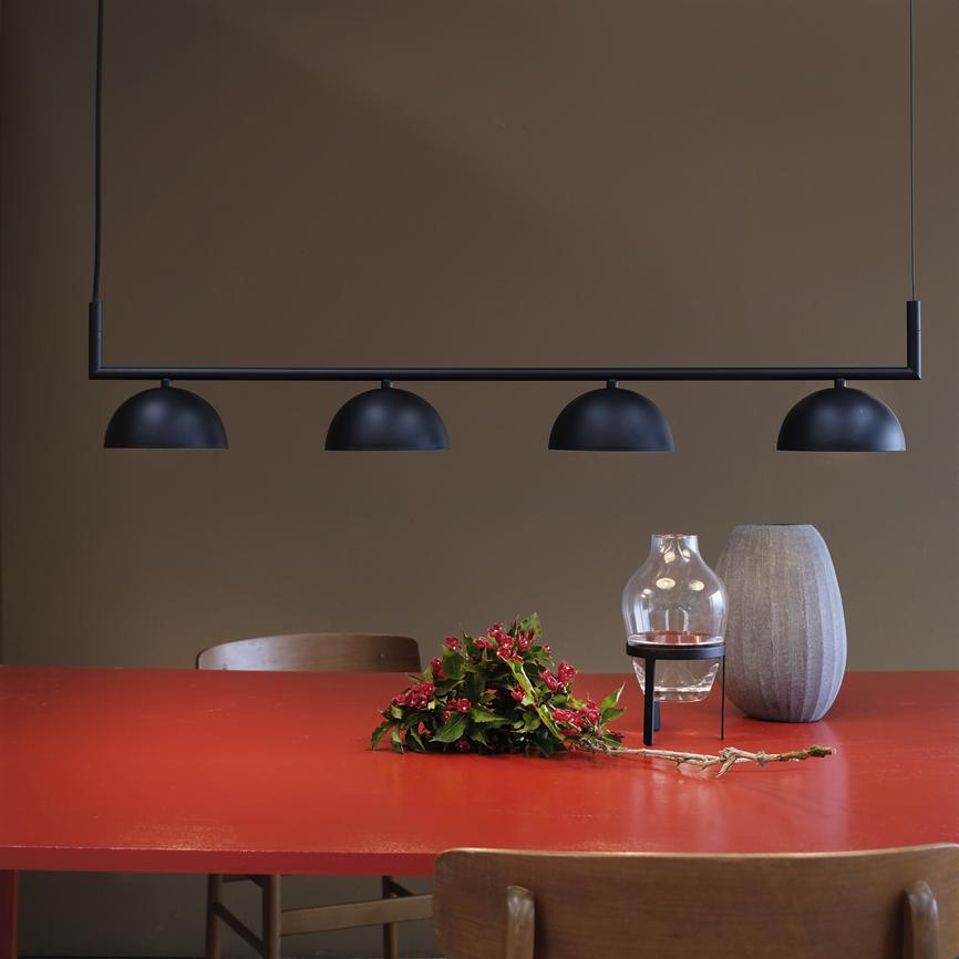 Fire skålformede, sorte loftlamper hænger på en stang over et rødt bord, hvor en lille buket, en glaslanterne og en grå keramisk vase står foran træstole mod en brun væg.