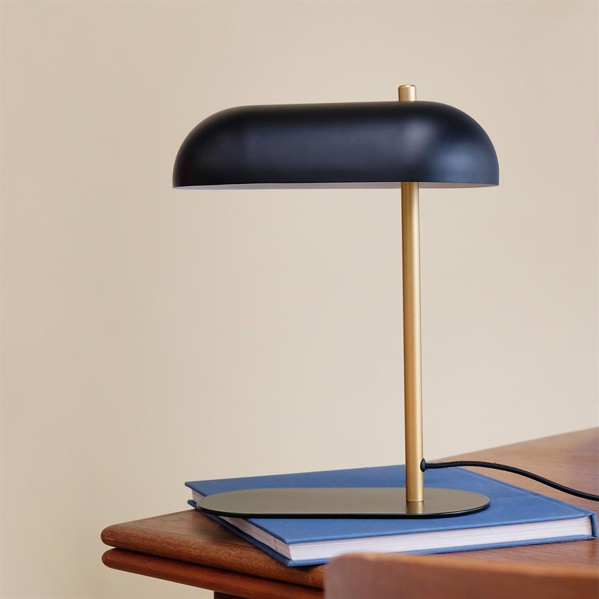 Bordlampe i sort skærm og guld stang står på en blå notesbog på et træbord foran en beige væg.