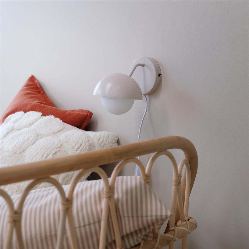 Væglampe monteret på væg, rettet mod sengen; en rund lampeskærm hænger over rattan-sengens gavl, omgivet af en creme pelsplaid og en rustfarvet pude.